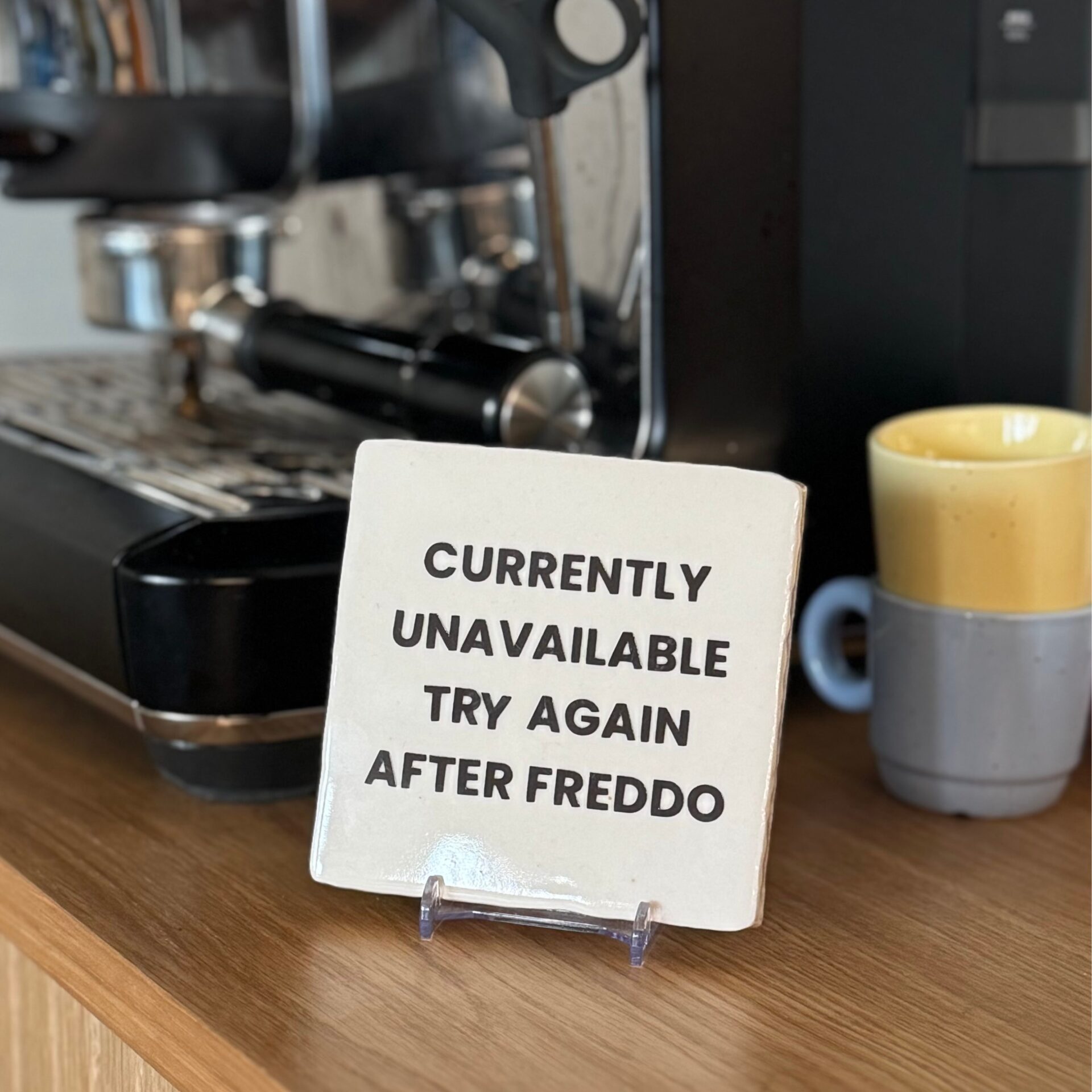 Wit tegeltje met de tekst Currently unavailable try again after freddo, op een standaard bij de koffiemachine