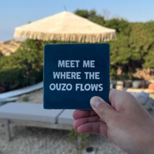 Donkerblauw tegeltje met de tekst Meet me where the ouzo flows, in de hand op Karpathos